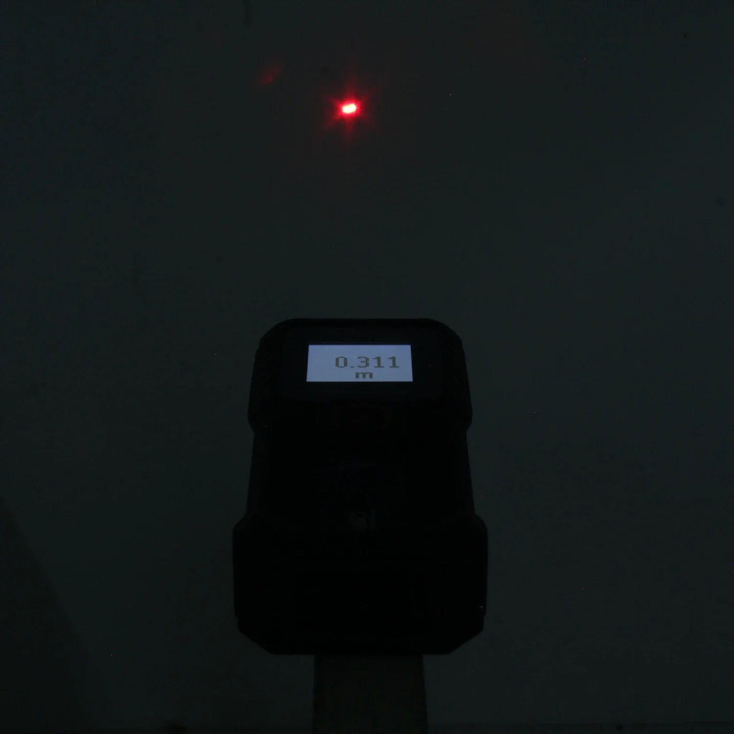 Medidor láser digital proyectando punto rojo, mostrando 0.311m en pantalla para medición de distancia.